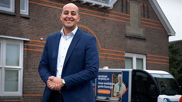 Foto van eigenaar Hamid Hamid Gongai van apotheek de Glimlach. Hij staat buiten en achter hem zie je een elektrische bestelbus
